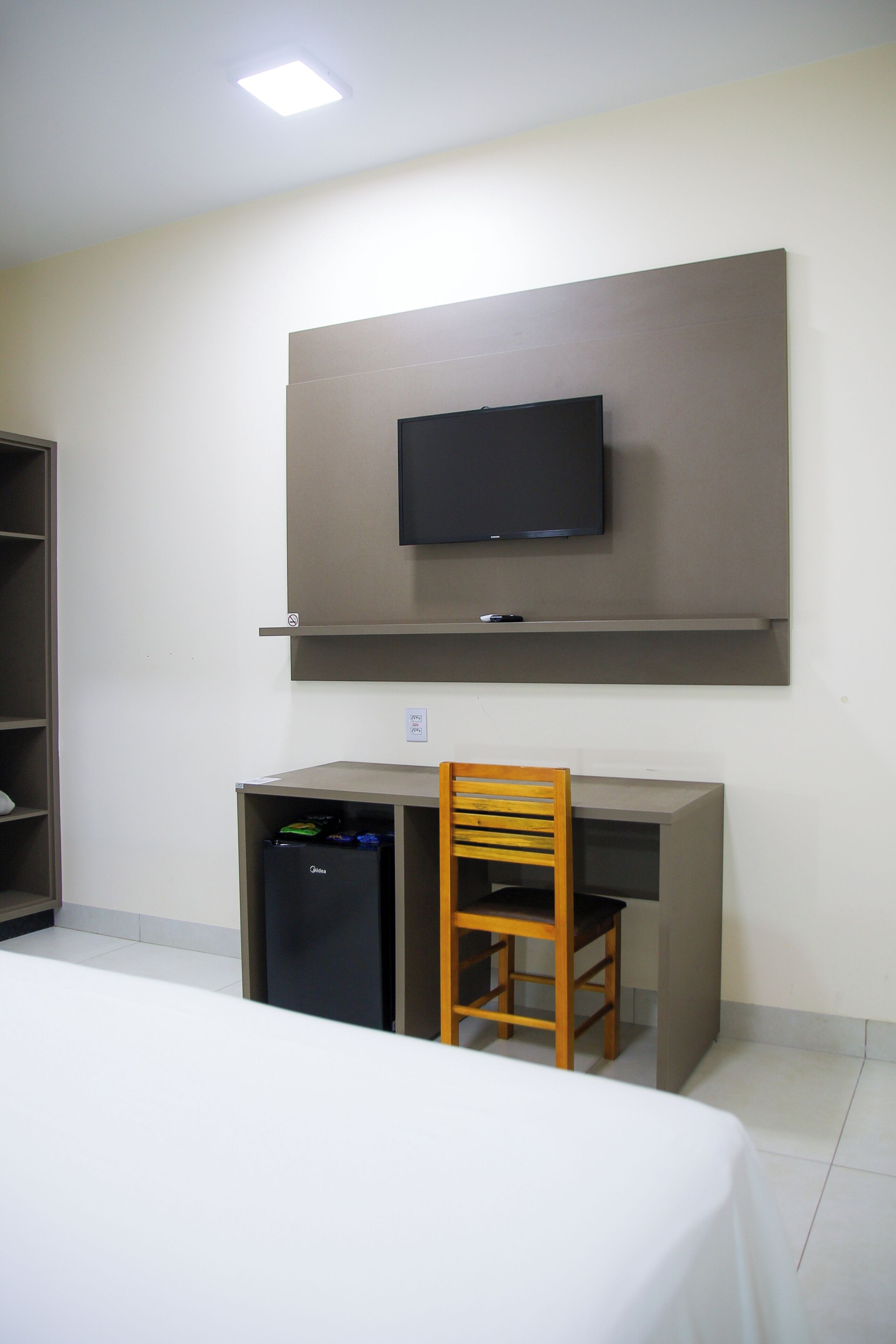 Une chambre comprenant un lit, un bureau, une chaise, un téléviseur fixé au mur et un petit réfrigérateur.