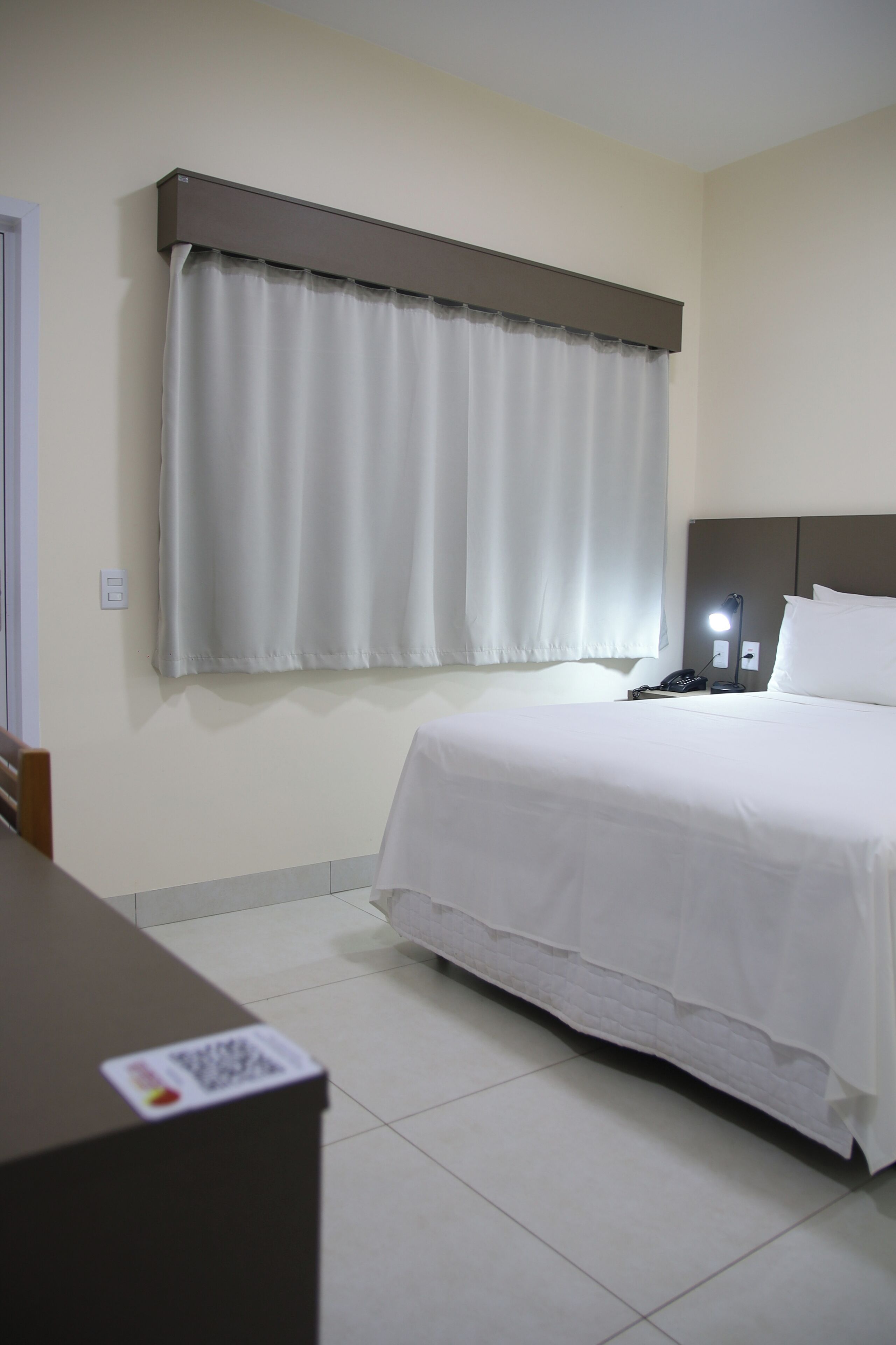 A hotel room with a bed, a bedside table, a lamp, and a window with curtains.