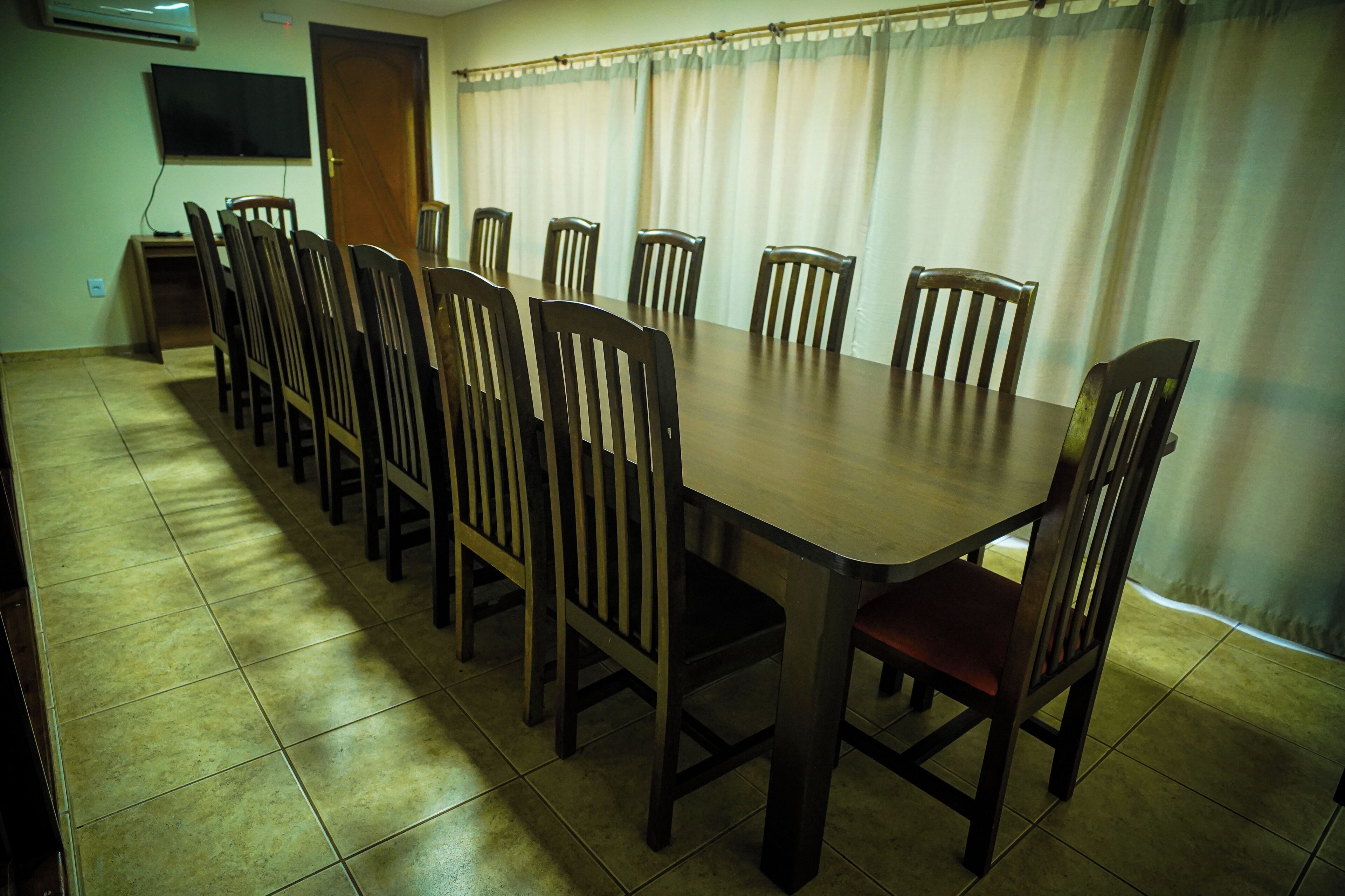 Un salón de conferencias con una larga mesa y múltiples sillas, un televisor montado en la pared y una puerta.