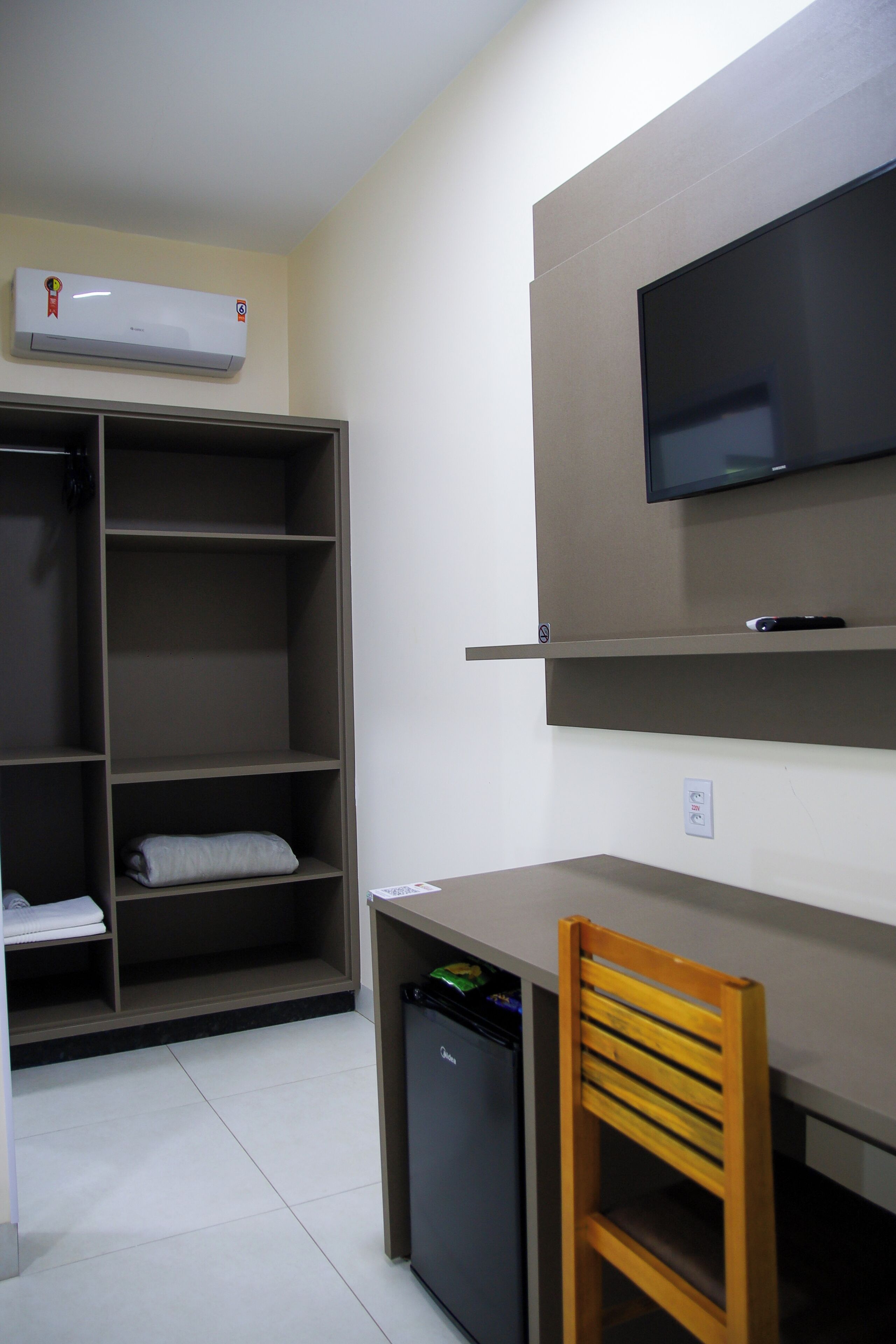 A room with a desk, a television mounted on the wall, a wardrobe with shelves, and a small refrigerator.