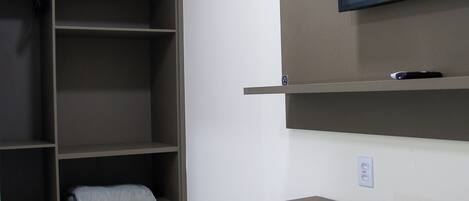 A room with a desk, a television mounted on the wall, a wardrobe with shelves, and a small refrigerator.