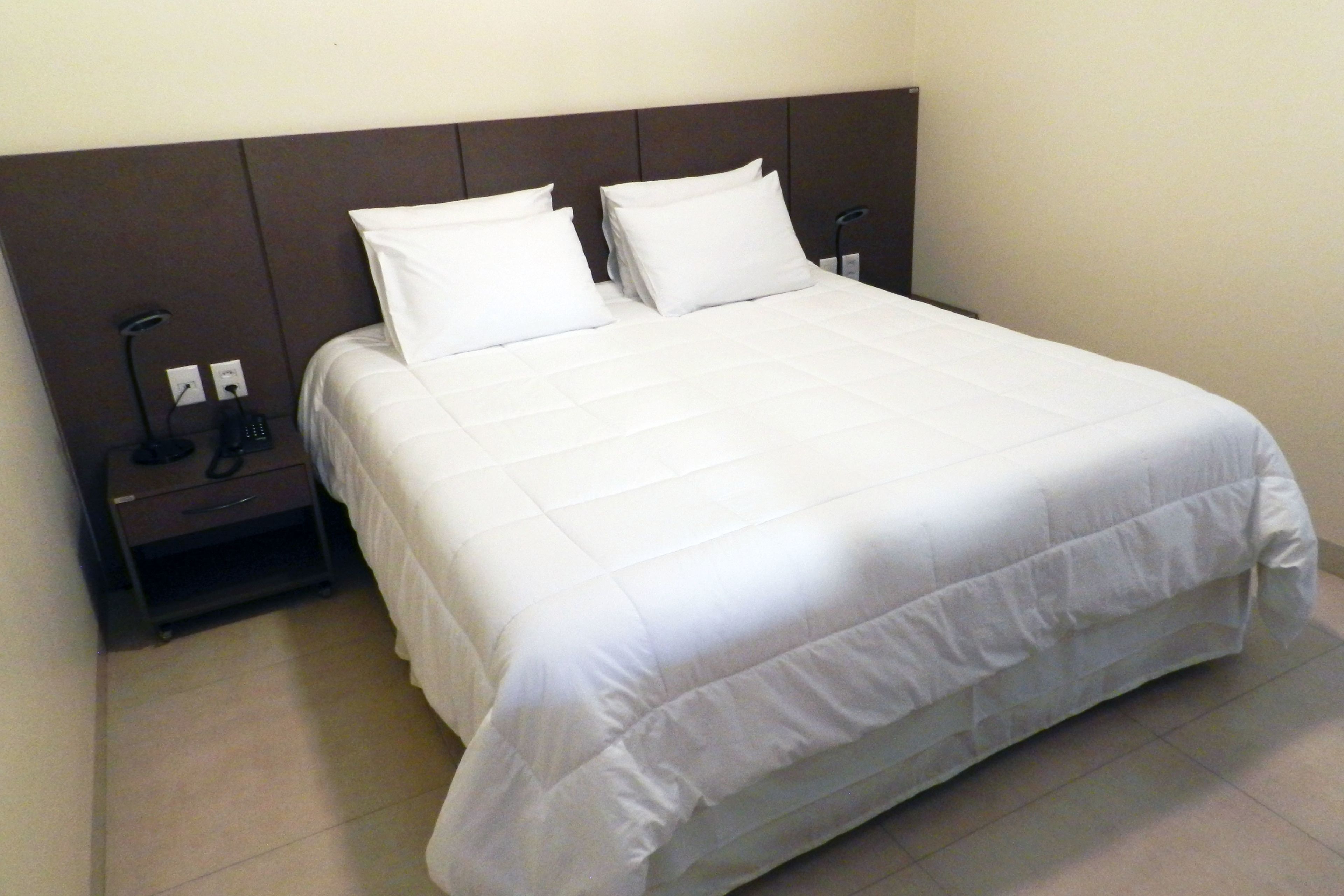 A neatly made bed with white bedding and a headboard, a bedside table with a lamp and a phone, and a power outlet on the wall.