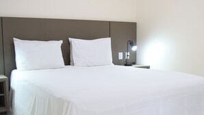 Uma cama bem arrumada com lençóis brancos, uma mesa de cabeceira com abajur e uma luminária na parede.