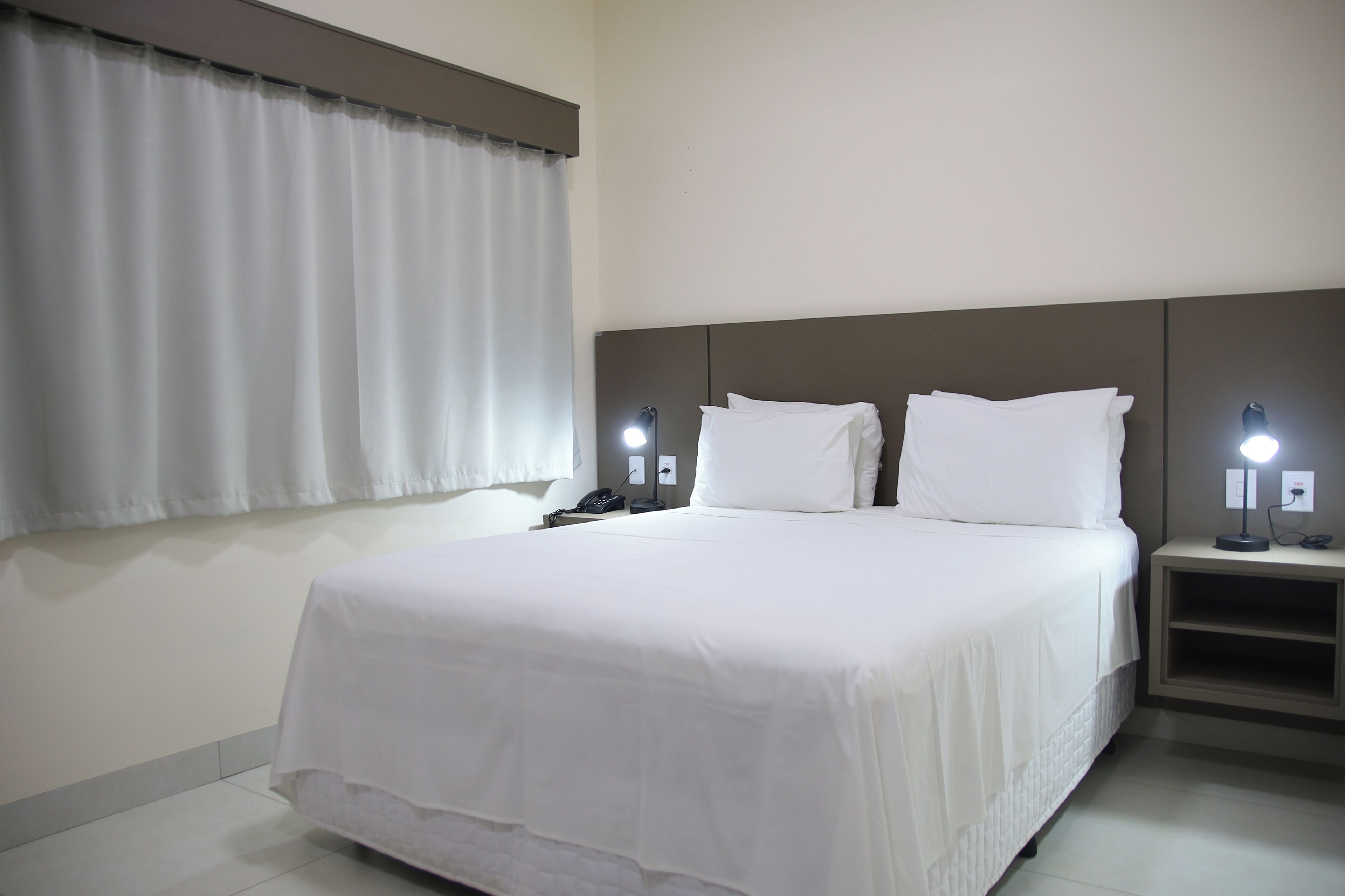 Ein Hotelzimmer mit einem großen Bett, zwei Nachttischen mit Lampen, einem Fenster mit Vorhängen und einem an der Wand befestigten Telefon.