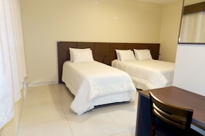 Two single beds with white linens and wooden headboards, a small table and chair, and a window with white curtains.