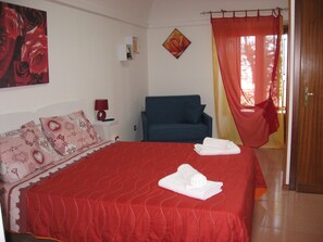 A neatly made bed with red bedding and white towels, a blue armchair, a small bedside table with a lamp, and a painting of roses on the wall. - San Procopio (Troina)