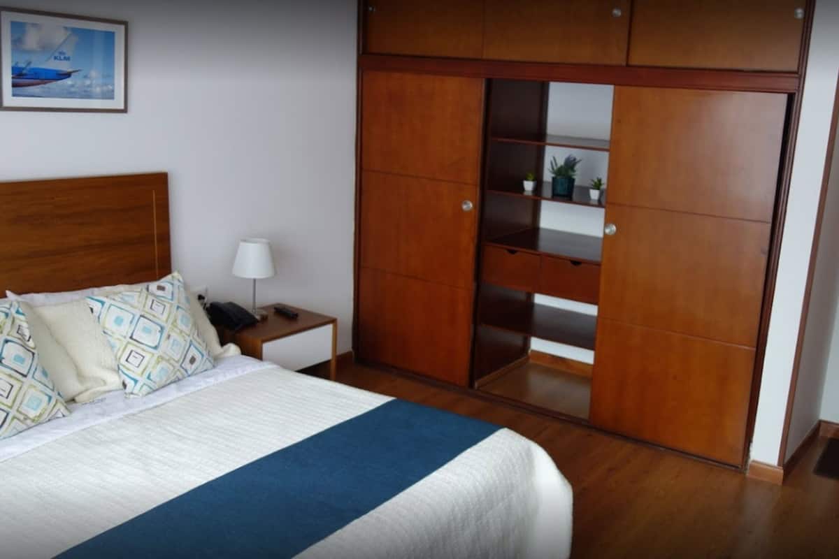 A bedroom with a bed, bedside table, lamp, wardrobe, and a bathroom visible through a glass door.