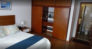 A bedroom with a bed, bedside table, lamp, wardrobe, and a bathroom visible through a glass door. - Hotel Maceo Chico (Bogotá)