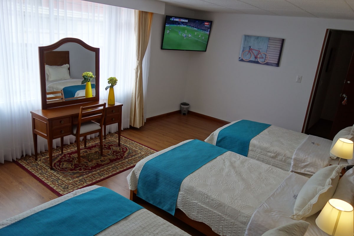 A hotel room with two beds, a desk with a chair, a mirror, a TV on the wall, and a painting.