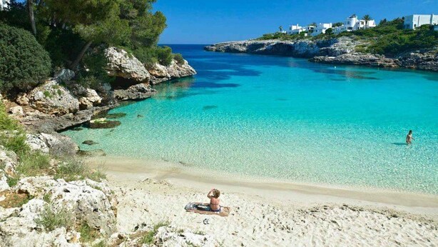 Une plage à l’eau turquoise et limpide, avec des affleurements rocheux et des bâtiments blancs perchés sur une falaise.
