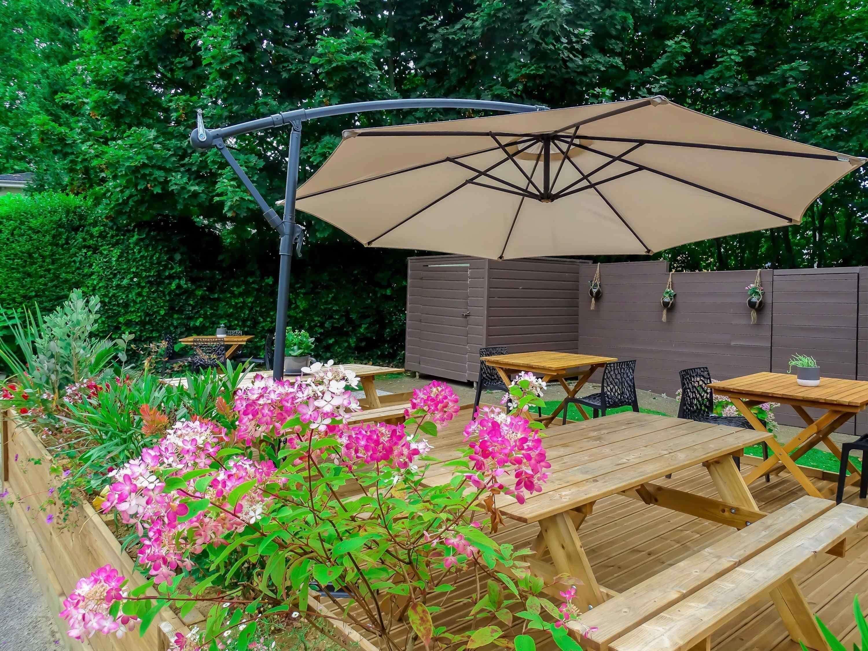 Un patio in legno con tavoli da picnic e un ombrellone, circondato da piante fiorite e una recinzione in legno.