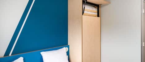 Uma cama com roupa de cama e almofadas brancas, um guarda-roupa de madeira com prateleira suspensa e uma parede de destaque azul.
