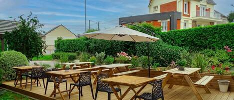 Un patio al aire libre con mesas y sillas de madera, rodeado de follaje y una casa al fondo.