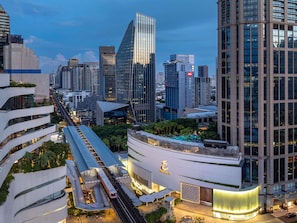 A modern hotel with a prominent glass facade, a rooftop pool, and a train station nearby.