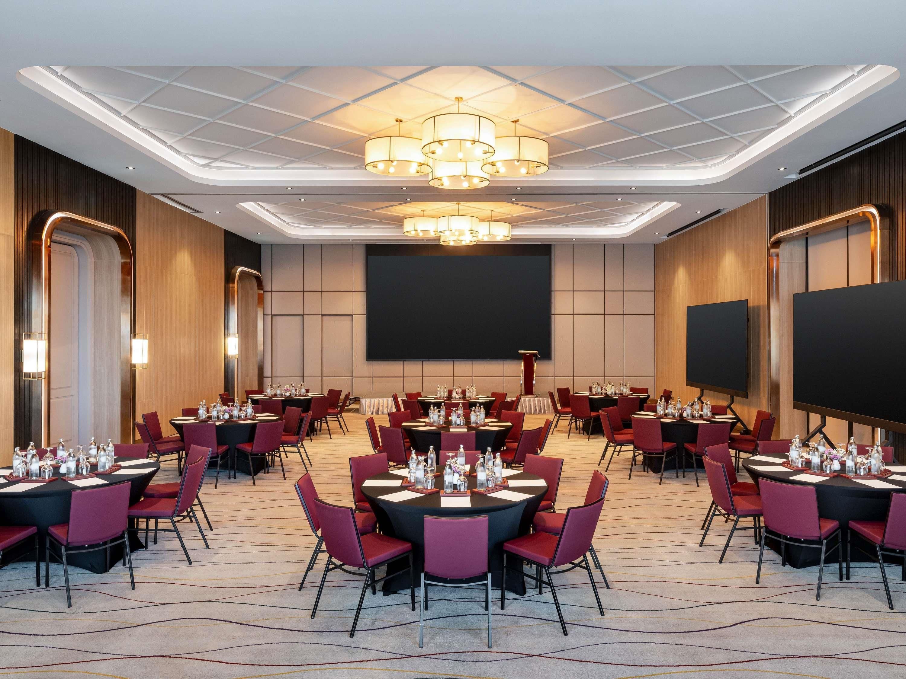 A conference room with multiple tables, chairs, a large screen, and a chandelier.