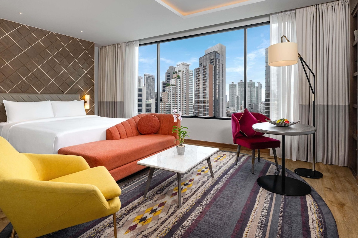 Ein Hotelzimmer mit einem Bett, einem Schlafsofa, einem gelben Sessel, einem kleinen Tisch, einer Stehlampe und Blick auf die Stadtlandschaft.