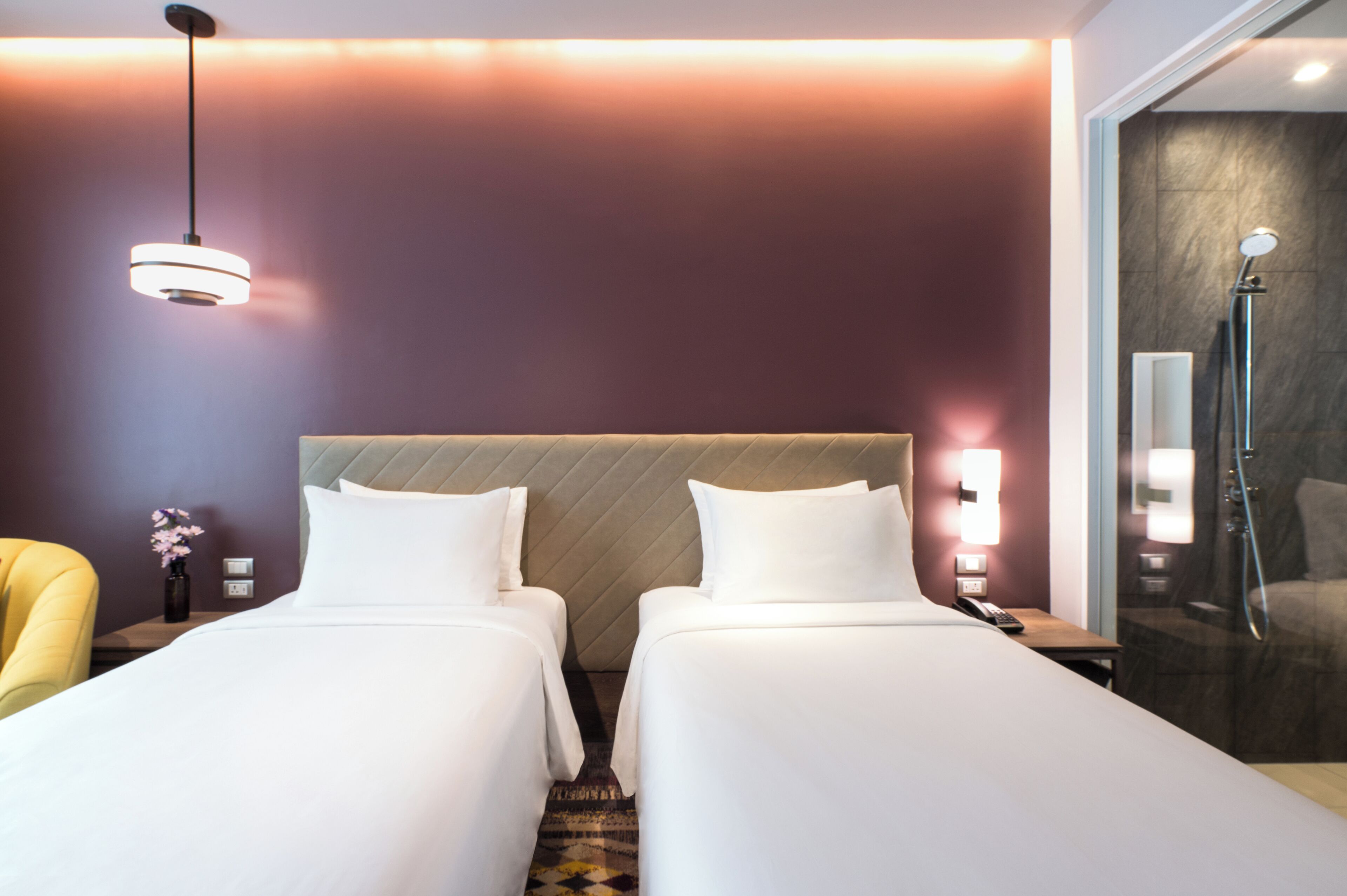 A hotel room with two beds, a nightstand, a chair, a vase of flowers, and a shower area.