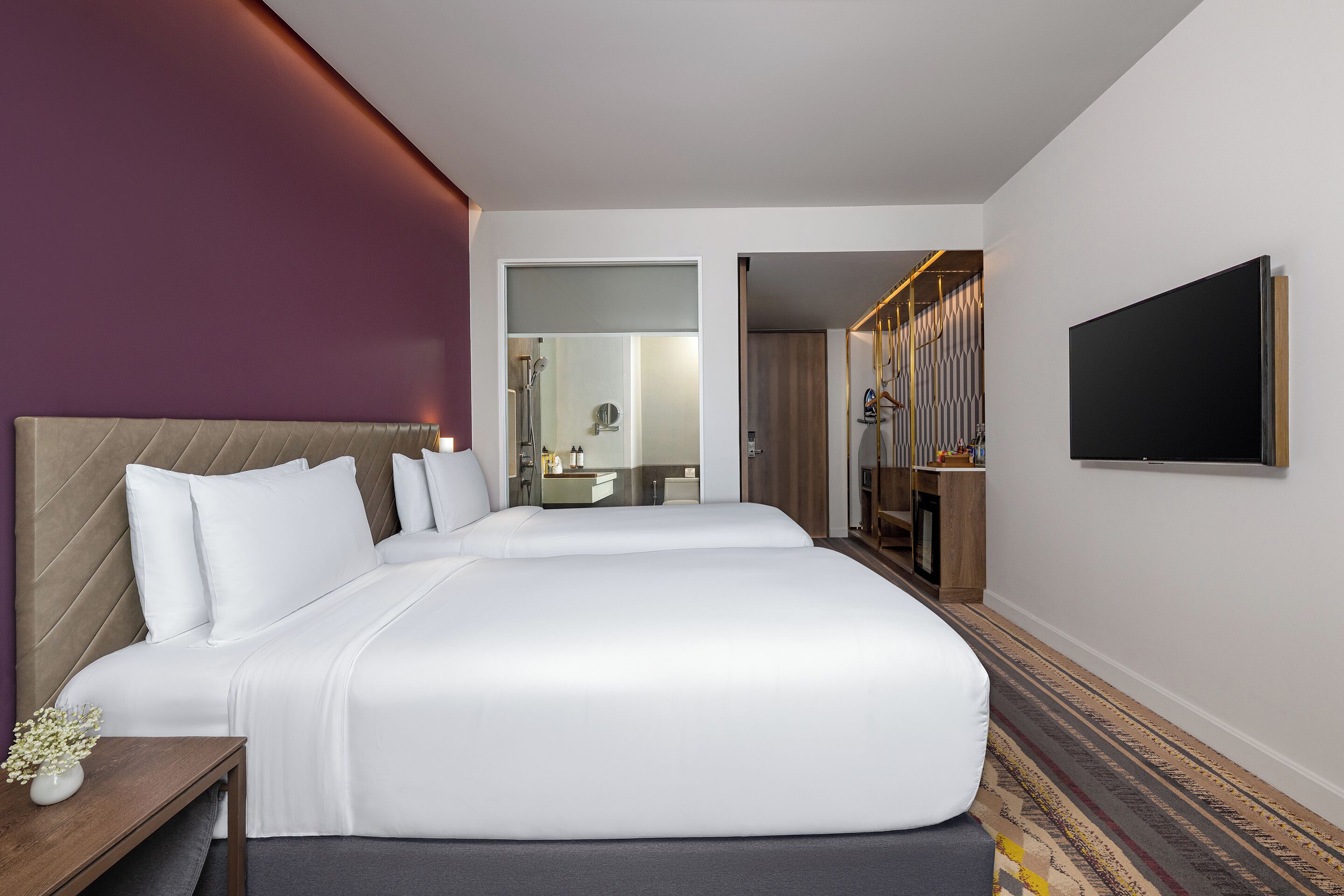 Ein Hotelzimmer mit einem großen Bett, einem Nachttisch mit einer Vase, einem Spiegel und einem an der Wand befestigten Fernseher.