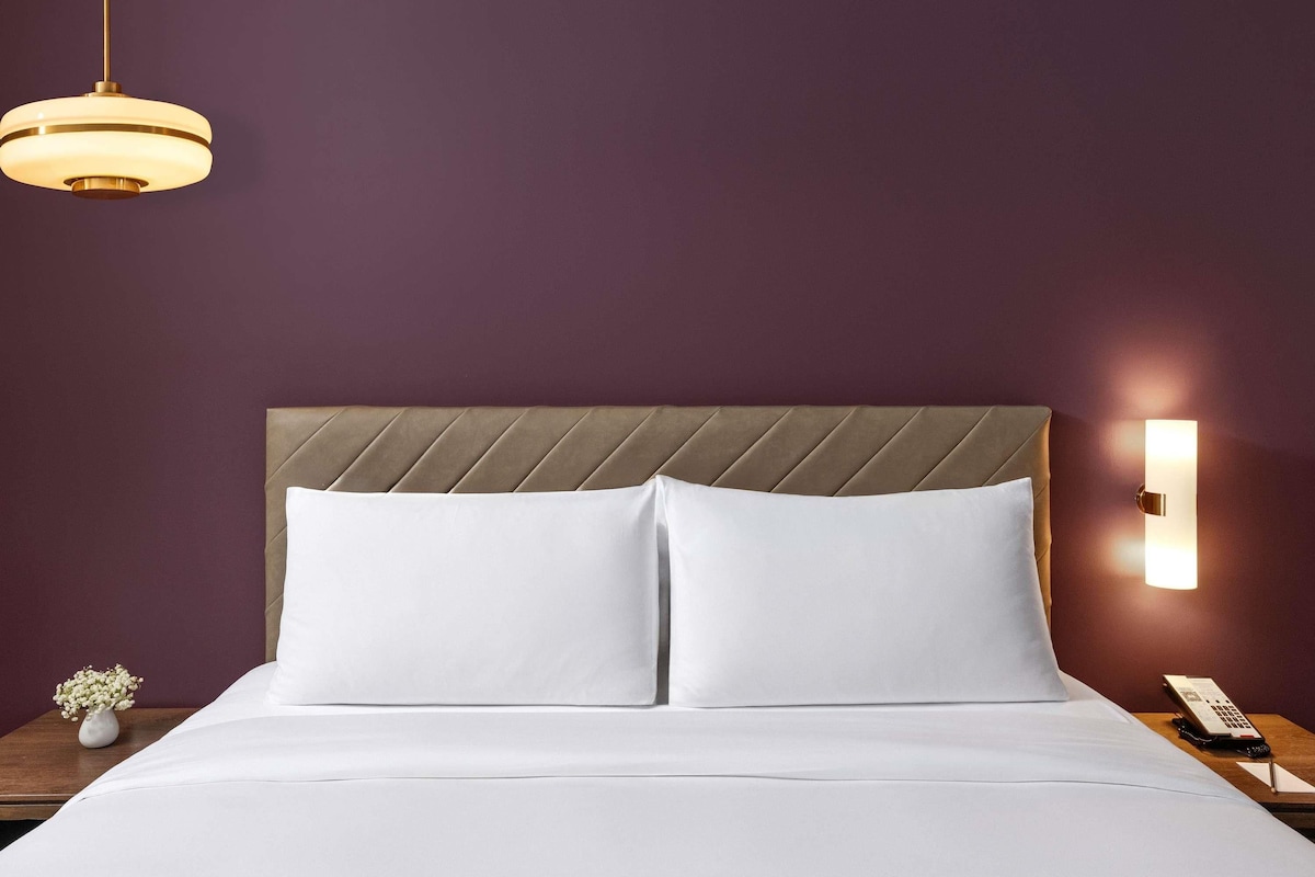 A hotel room with a bed, a nightstand, a vase with flowers, a wall-mounted lamp, and a telephone.
