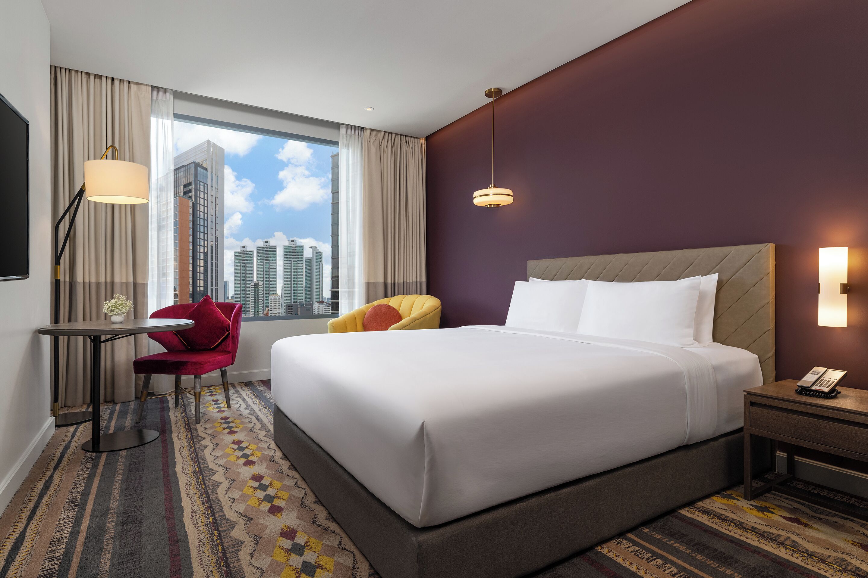 A hotel room with a large bed, a desk with a chair, a window with curtains, a city view, and a patterned carpet.