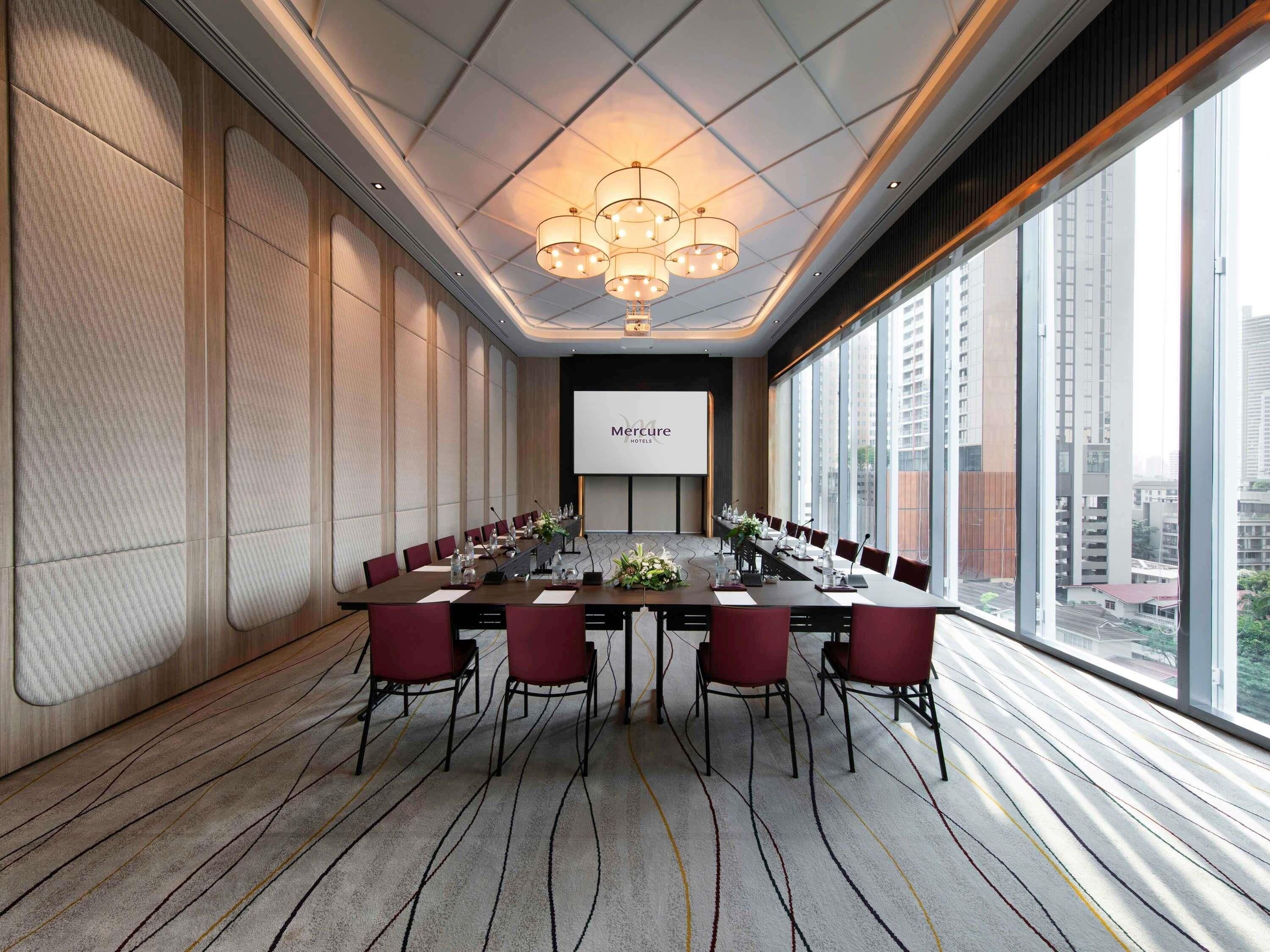 A conference room with a long table, red chairs, a projector screen, and large windows offering a city view.