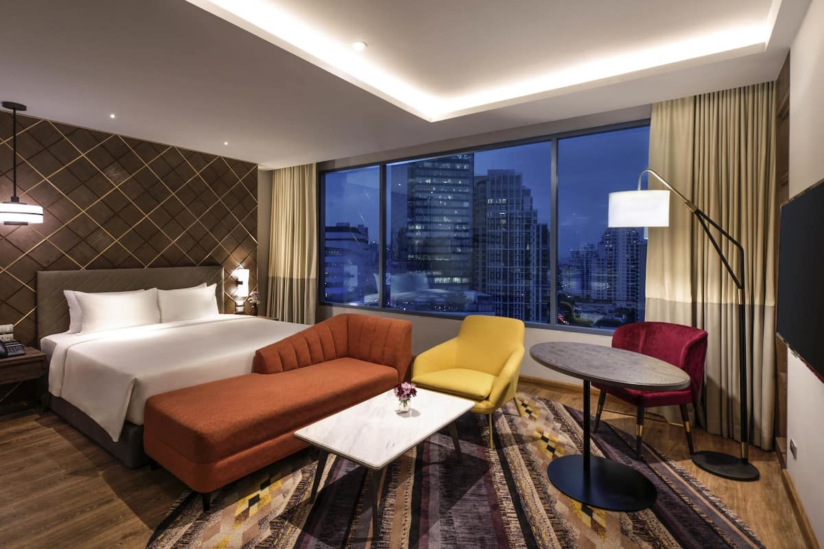 A hotel room with a large bed, a sofa, a small table, a chair, a floor lamp, and a view of the cityscape.