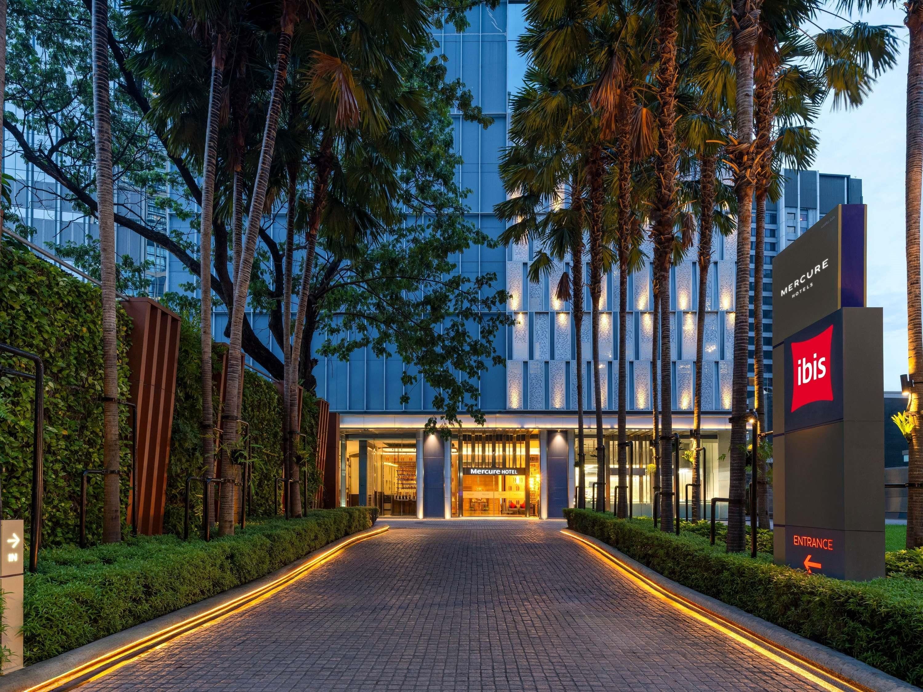 Una entrada de hotel con un sendero pavimentado que lleva a las puertas, rodeada de altas palmeras y un edificio moderno al fondo.