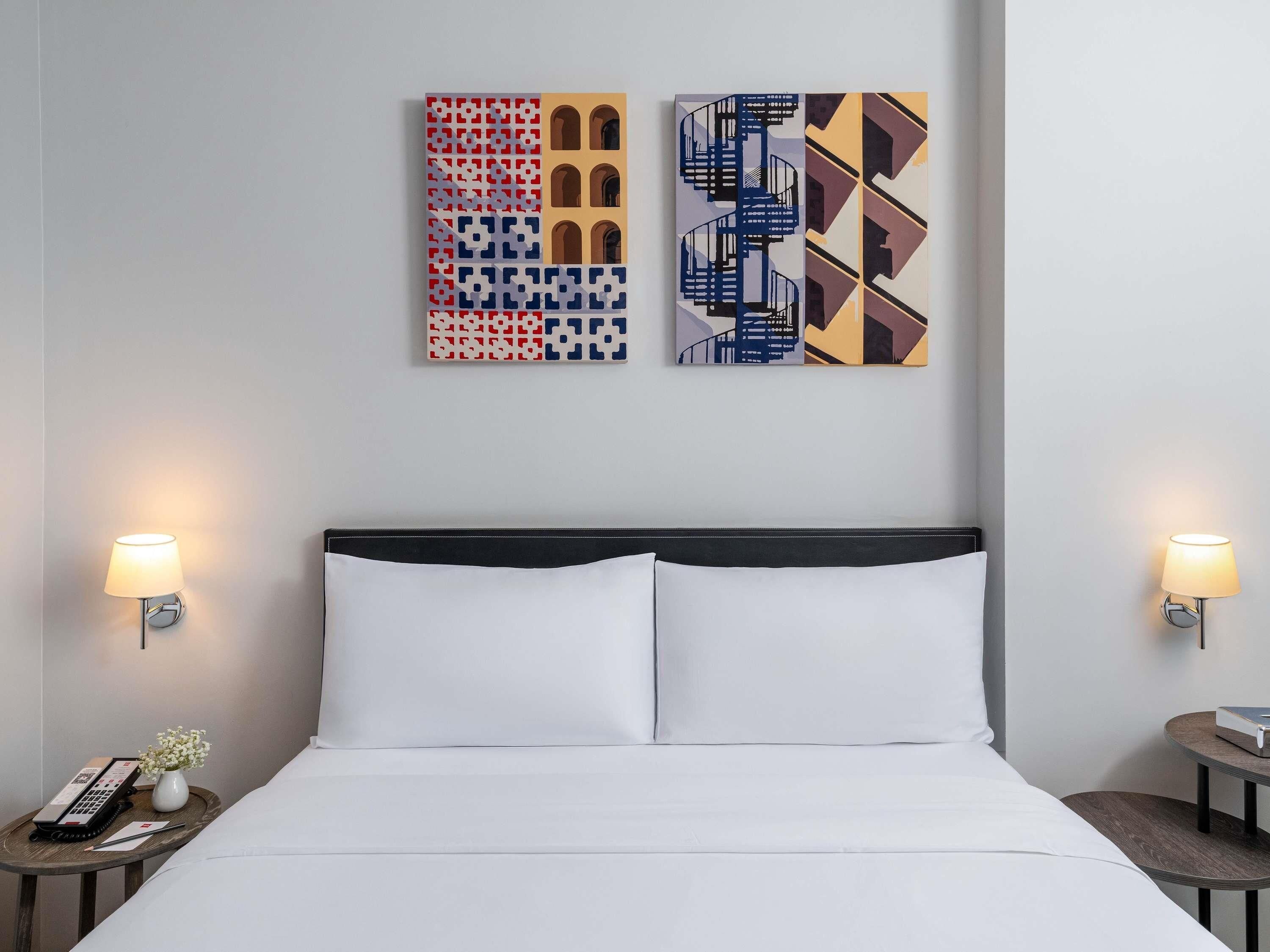 A hotel room with a bed, two bedside tables, a wall-mounted lamp, and two pieces of wall art.