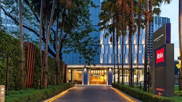 A hotel entrance with a paved pathway leading to the doors, surrounded by tall palm trees and a modern building in the background.