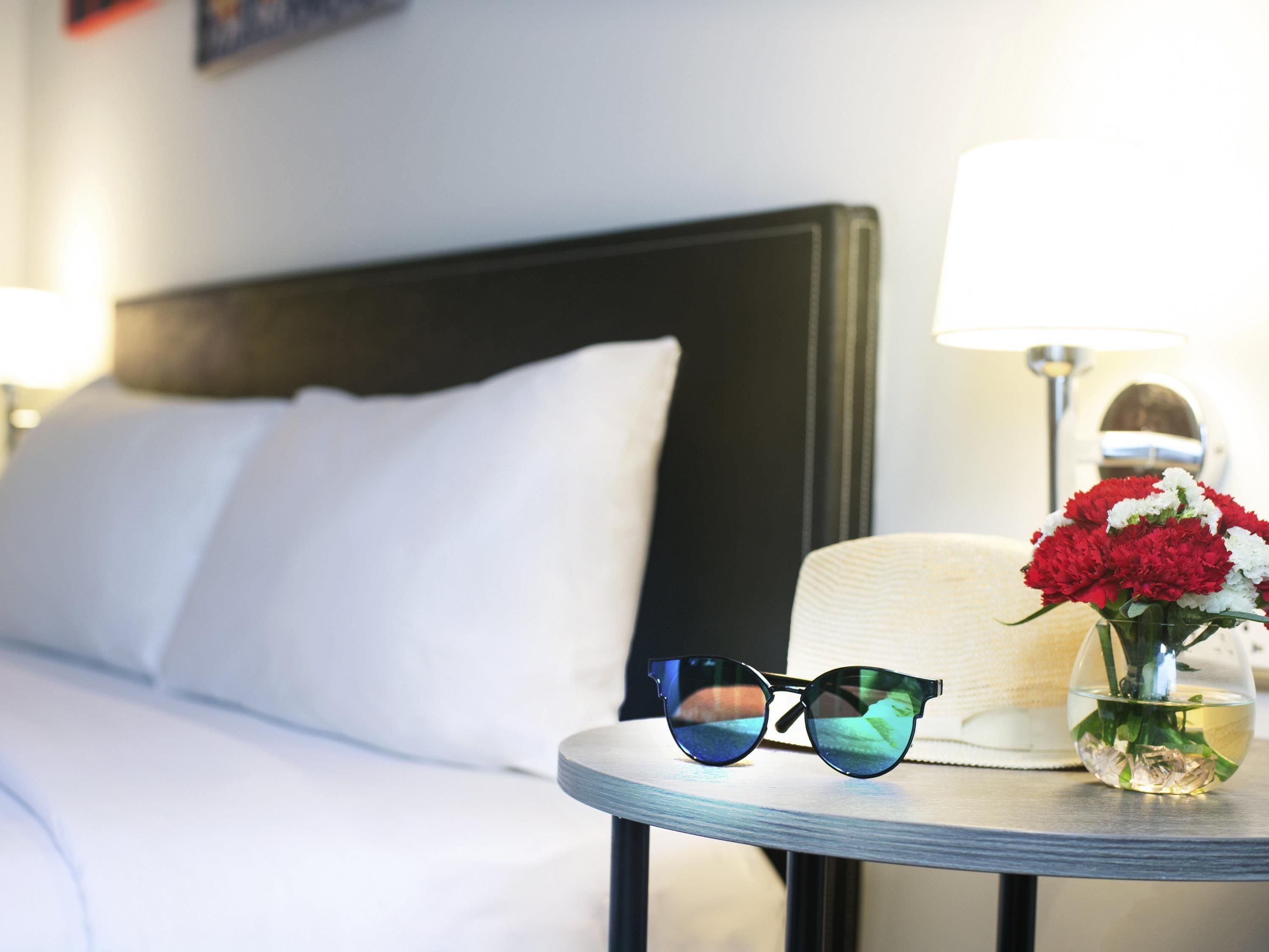 A neatly made bed with white linens, a round table with a lamp, and a vase with red and white flowers.