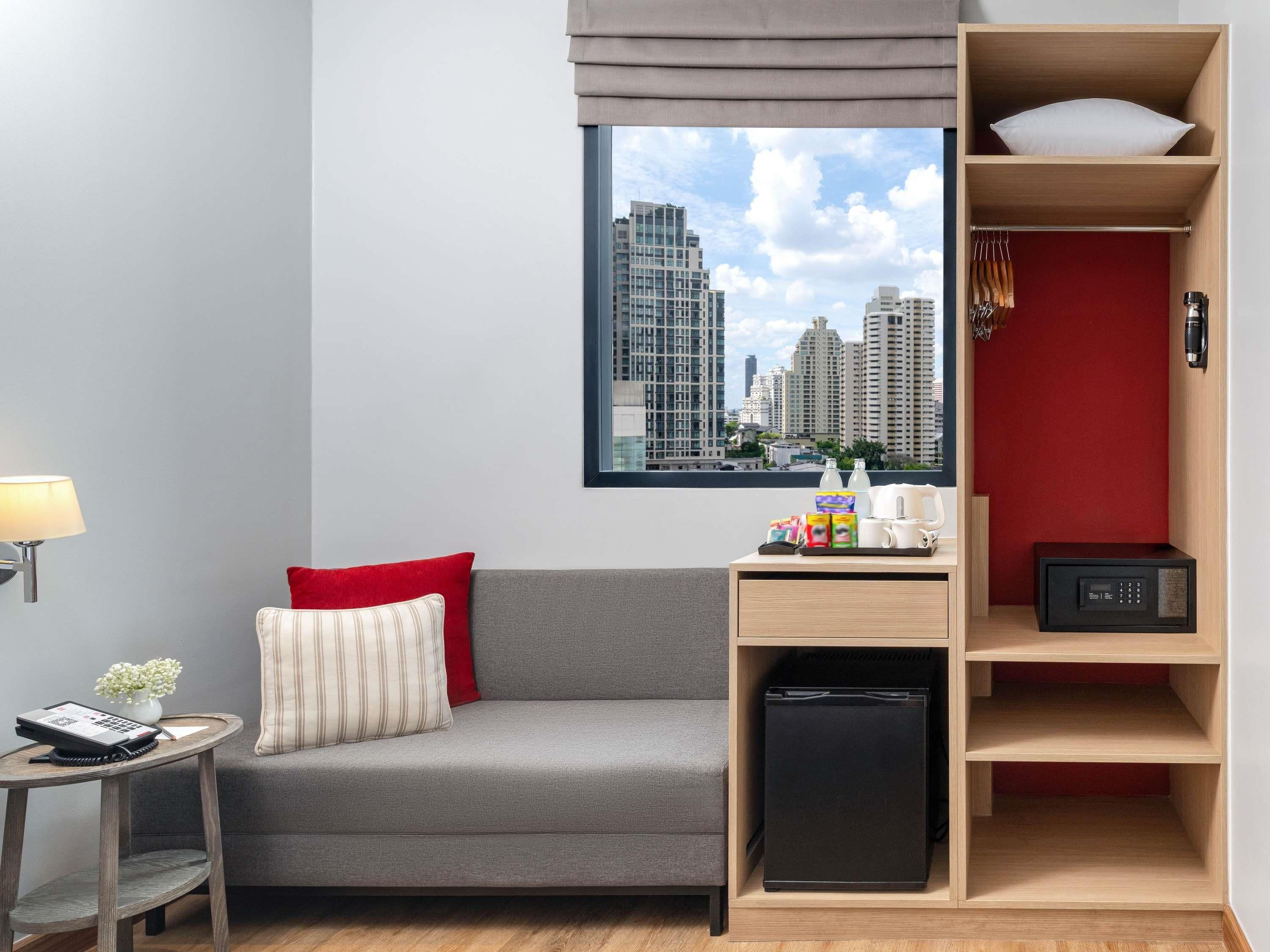 A modern hotel room with a grey sofa, a small table with a lamp, a red wall, a window with a city view, and a wooden shelf with a kettle and a small fridge.