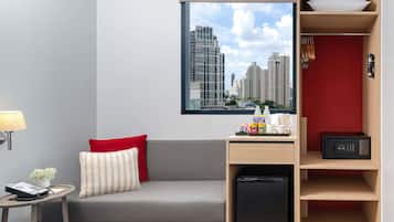 A modern hotel room with a grey sofa, a small table with a lamp, a red wall, a window with a city view, and a wooden shelf with a kettle and a small fridge.