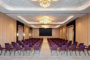 Un salón de conferencias con filas de sillas moradas, una pantalla grande y una lámpara de araña moderna.