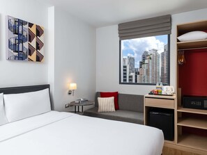 A hotel room with a bed, a seating area, a desk with a computer, a wardrobe, and a window with a cityscape view. - Ibis Bangkok Sukhumvit 24 (Bangkok)