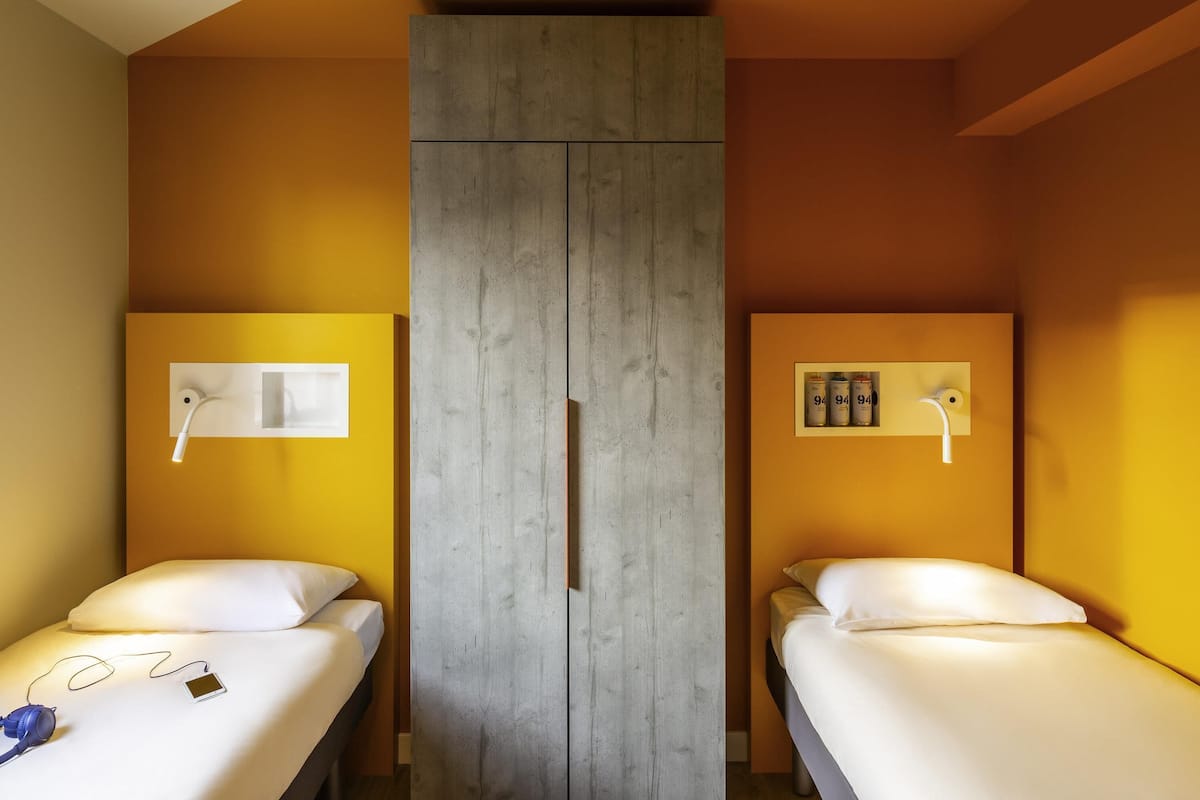 A room with two beds, a wardrobe, and a wall-mounted shelf with cans.