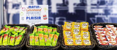 A display of assorted packaged goods in baskets, including items labeled 'MEL - HONEY' and 'MEL - HONEY + JEL,' with a sign promoting 'MATIN - Gourmand' and 'LA TOUCHE PLAISIR.'