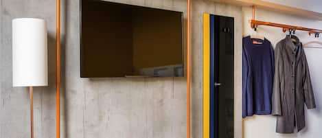 Una habitación moderna con un televisor montado, una estantería con un control remoto y un perchero con ropa.