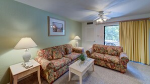 A living room with two floral-patterned sofas, a small table with a lamp, a framed picture on the wall, and a ceiling fan.