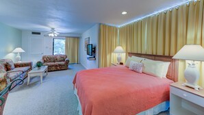 A hotel room with a large bed, two armchairs, a small table, a TV, and a ceiling fan.