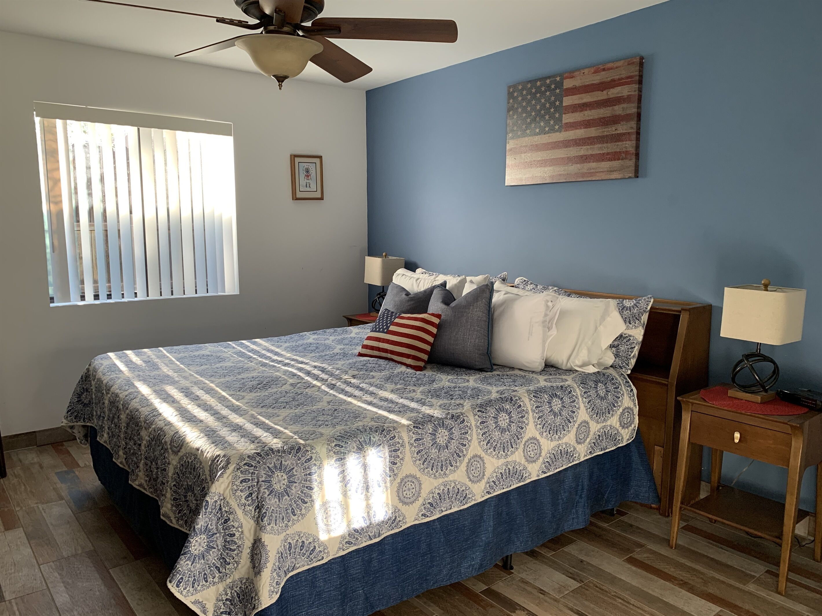Een slaapkamer met een bed, een plafondventilator, een nachtkastje met een lamp, een raam met jaloezieën en een Amerikaanse vlag aan de muur.