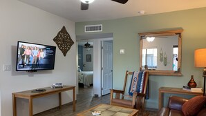 Un salon comprenant un téléviseur, une table basse, un canapé et un fauteuil. Une porte donne accès à une autre pièce.