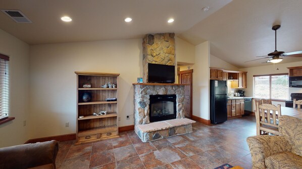 A spacious living area with a stone fireplace, wooden shelves, and a kitchen with wooden cabinets and a black refrigerator.
