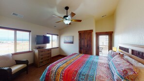 A bedroom with a large bed, a ceiling fan, a TV, and a view of the outdoors through the windows.