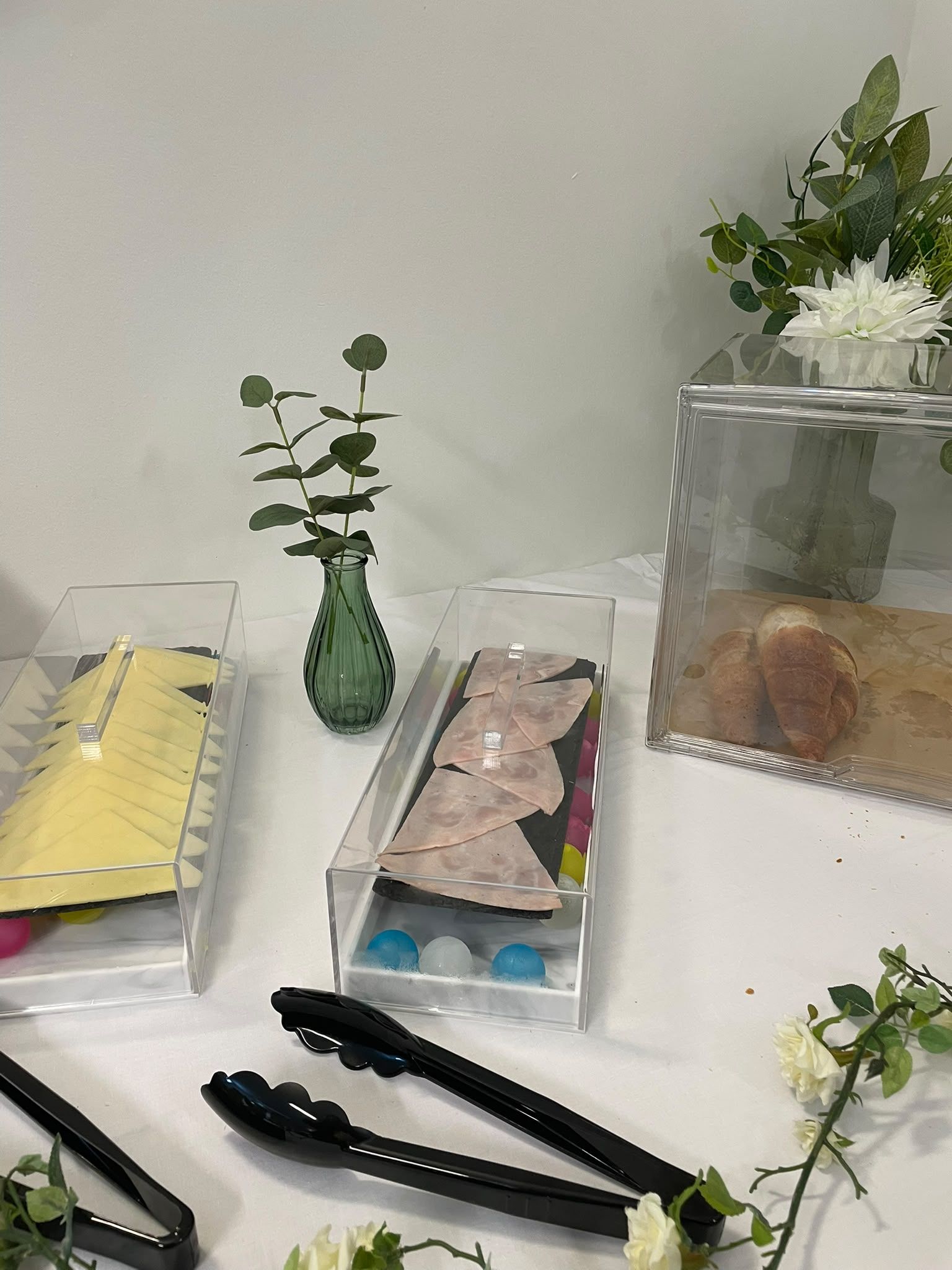 A table with various items including a vase with a plant, a container with a white flower, a box with a yellow object, a box with blue and white objects, and a box with a brown object.