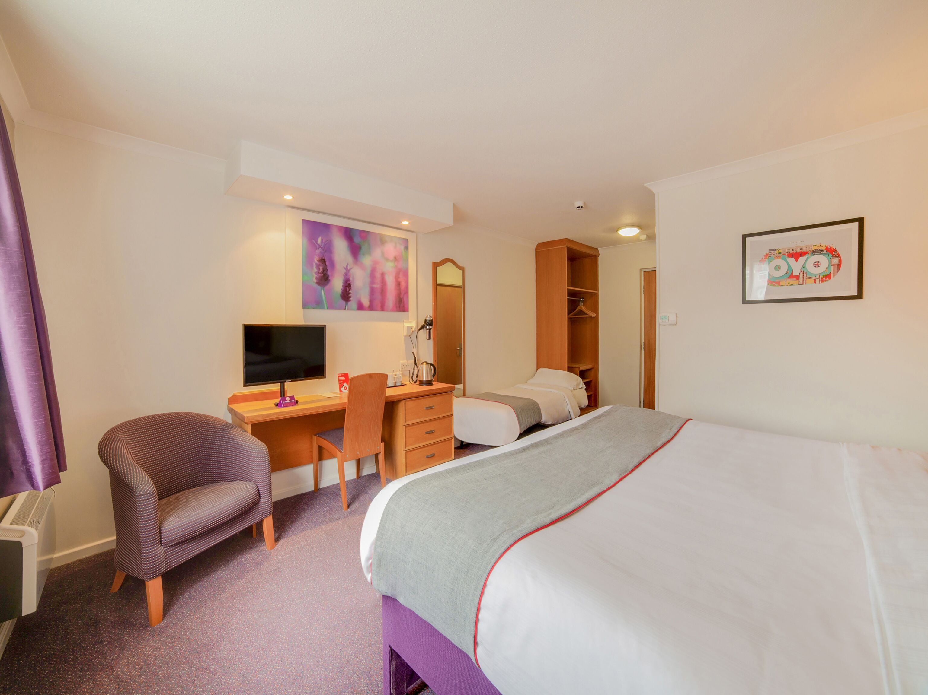 A hotel room with a bed, a desk with a computer, a chair, a wardrobe, and a framed picture on the wall.