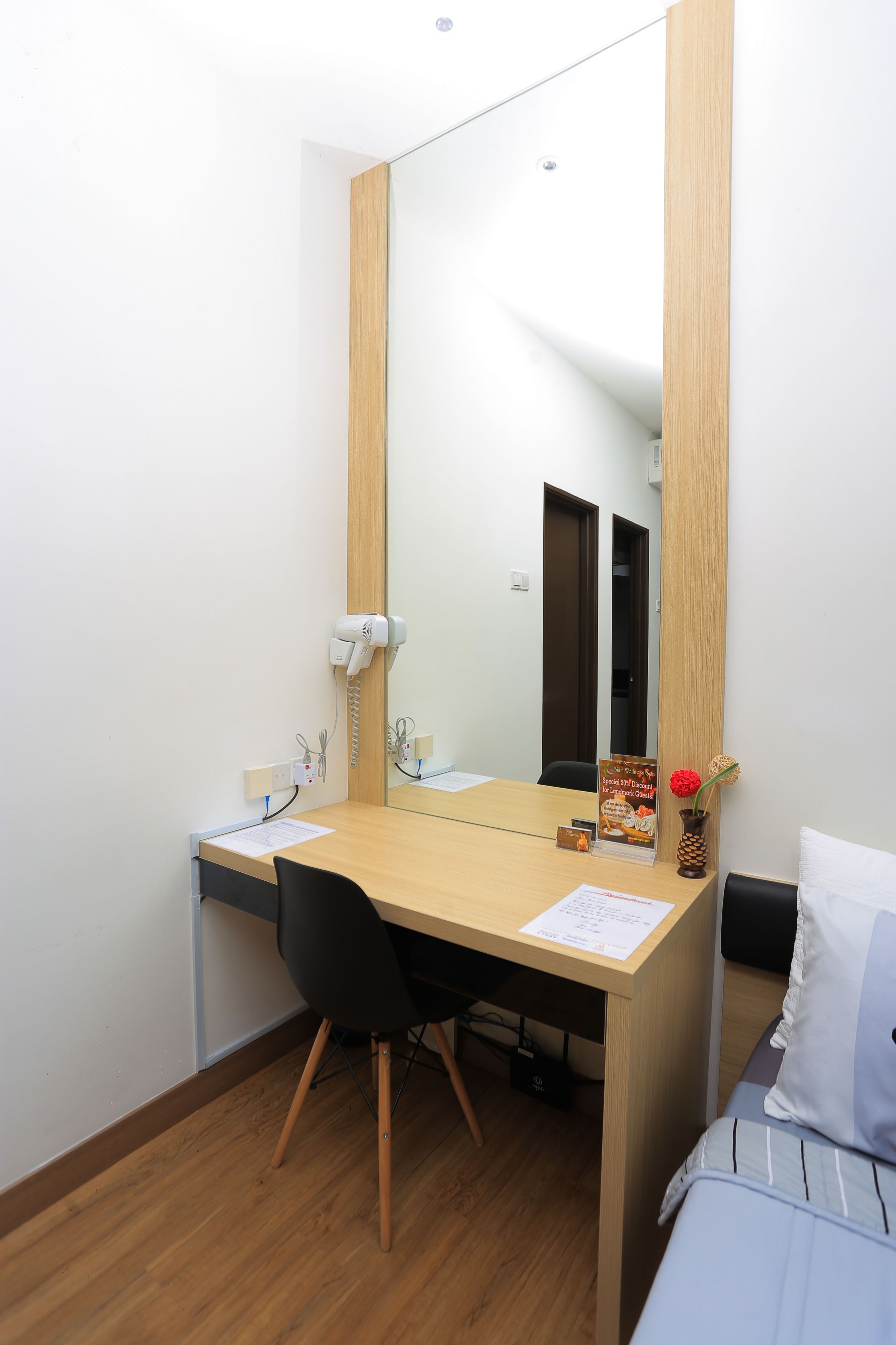 Quarto pequeno e moderno com escrivaninha de madeira, cadeira preta, cama com manta listrada, espelho grande e um vaso pequeno com uma flor vermelha.