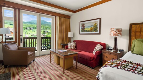 Una camera d'hotel con un divano rosso, una scrivania in legno, una sedia, un letto e vista su un paesaggio verde dalle finestre.