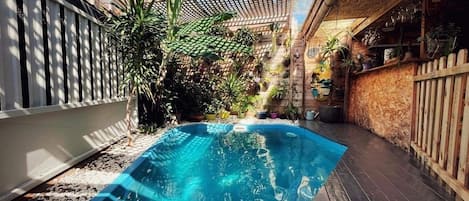 A small, enclosed swimming pool with a wooden deck surrounding it, surrounded by various plants and a wooden fence.