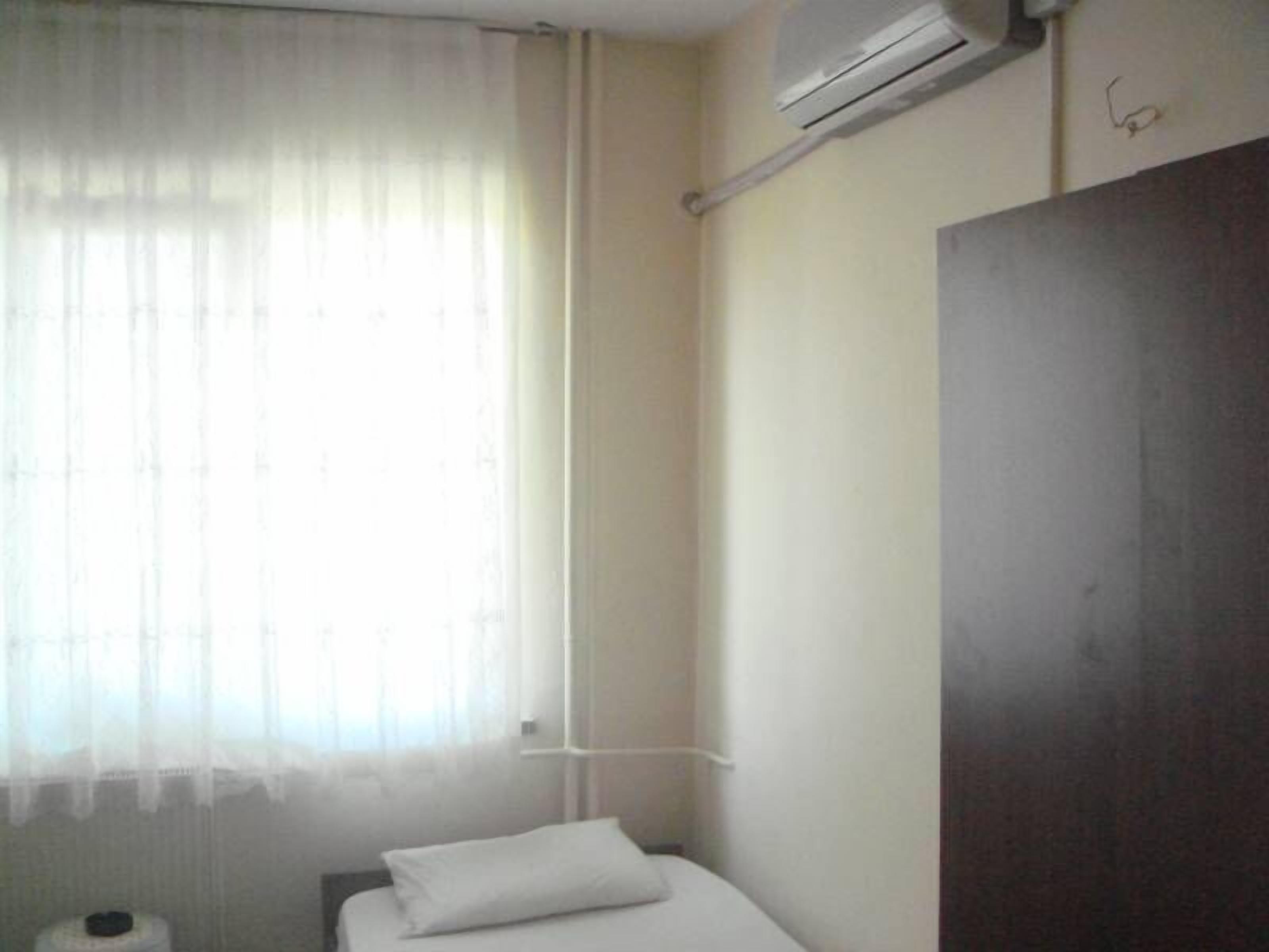 Une petite pièce simple, avec un lit, une fenêtre ornée de rideaux légers, un appareil de climatisation et une armoire en bois.