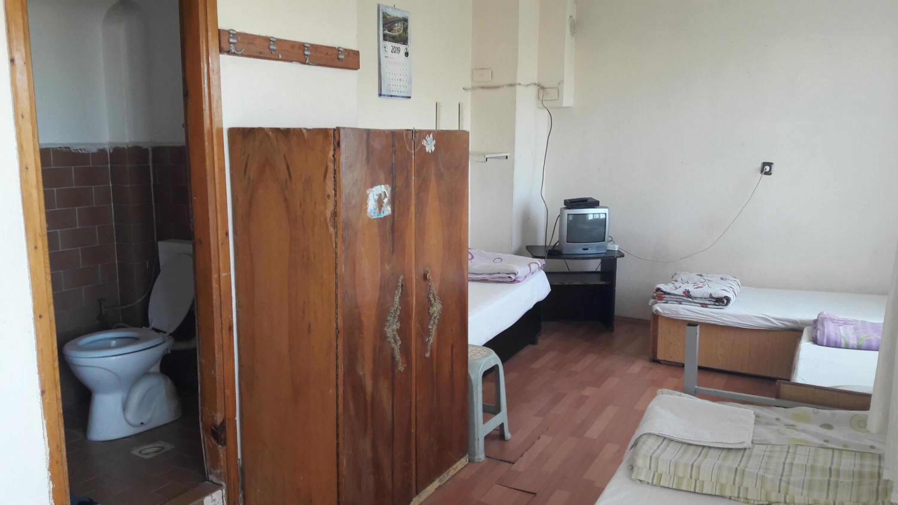 Quarto pequeno e compacto com divisória de madeira separando o espaço do toalete da área de dormir. Uma televisão está posicionada sobre uma mesinha ao lado da cama.